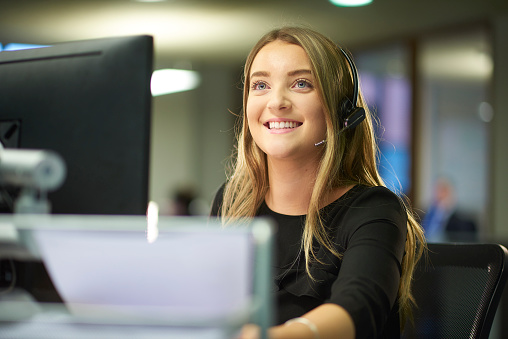 a teenage office worker sits at her desk and speaks on her telephone headset . She is smiling and looks happy to be working in a big open plan office .