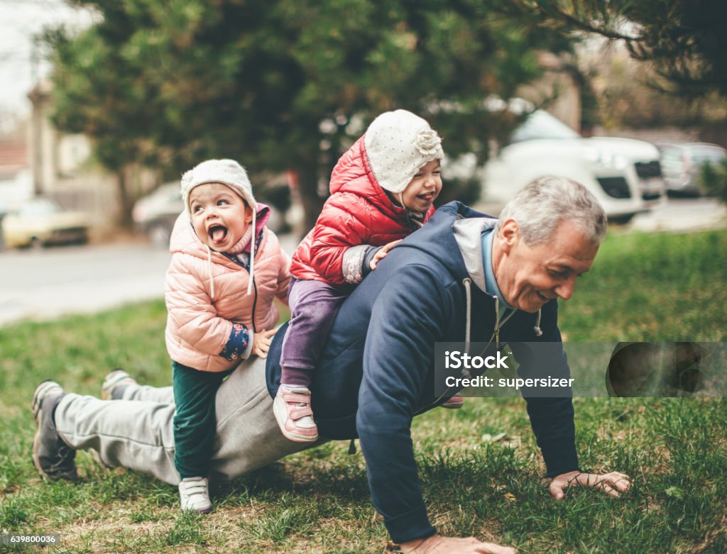 We exercise togather A photo of a playful grandfather and granddaughter. They are casually dressed and playing in the park. They exercise together. A grandfather is exercising while granddaughters are sitting on his back. Senior Adult Stock Photo