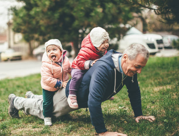 We exercise togather A photo of a playful grandfather and granddaughter. They are casually dressed and playing in the park. They exercise together. A grandfather is exercising while granddaughters are sitting on his back. push ups stock pictures, royalty-free photos & images