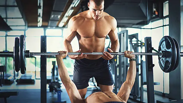 Closeup of teamwork at a gym. There are two guys, one of them is lifting the barbell and the other one is spotting him. Blurry gym equipment in background.