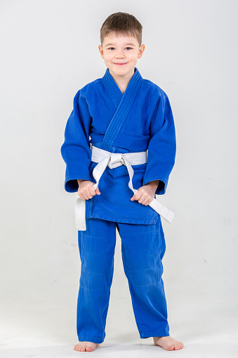 Cute elementary aged boy dressed in blue judo gi (sport uniform). The boy judo fighter is standing in judo position. He is smiling looking at the camera. Studio shooting on white background