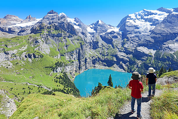 Children Hiking in the Swiss Mountains in Summer Two young kids are hiking in the Swiss Alps at Oeschinensee mountain lake on a beautiful summer day. The boys are walking on a narrow alpine trail overlooking a turquoise blue mountain lake below. lake oeschinensee stock pictures, royalty-free photos & images