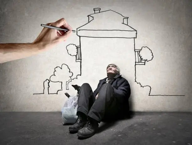 Homeless man is sitting on the ground while  hand is drawing a house on the wall
