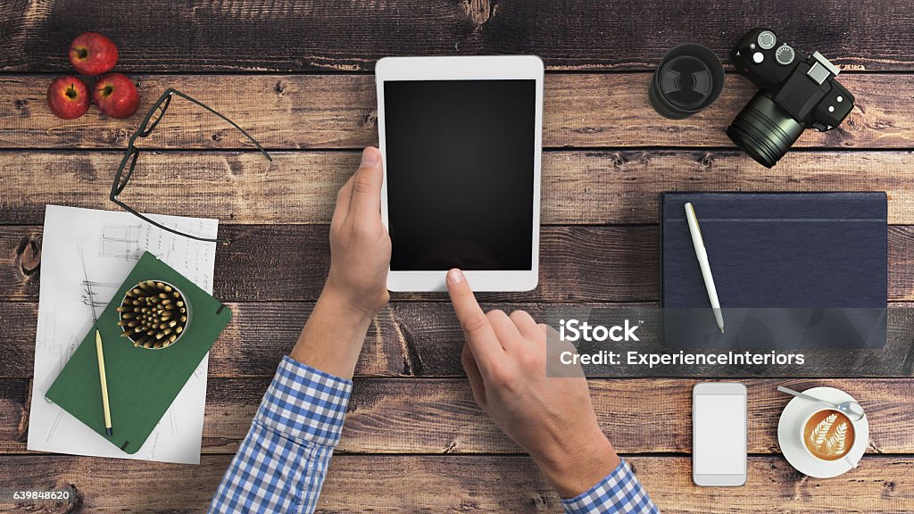 Man hipster holding tablet knolling overhead view Man holding blank tablet in hands. Knolling overhead office desk view, dark vintage old wood, with central part free for copy space. On the sides of the photo there are a notebook, a digital camera, coffee, smartphone, apples, glasses, pencils. Designer mockup. Horizontal composition. Desk Stock Photo