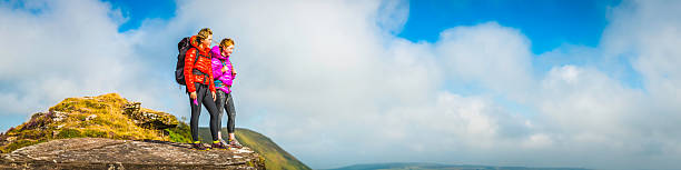 Teenage hikers in vibrant outdoor gear standing on a high mountain ridge looking out over a panoramic summer cloudscape.