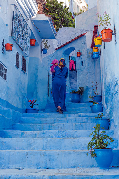 Woman in Blue Djellaba and Child in Blue City, Morocco Chefchaouen, Morocco. - April 12, 2014: A woman in a blue Moroccan cloak djellaba and with a girl in pink in her hands, is walking up the stairs in the blue painted Medina streets of Chefchaouen, Morocco. Chefchaouen is famous as most of the Medina streets are painted blue.North Africa,Nikon D3x moroccan girl stock pictures, royalty-free photos & images