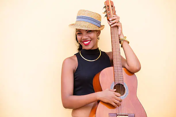 A portrait of a beautiful, young, smiling Cuban woman in Panama hat with a guitar in front of a yellow wall. Havana, Cuba, 50 megapixel image, copyspace.