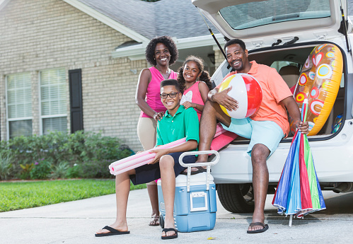 An African American family of four packing their car for a trip to the beach or pool, with beach ball, cooler, inflatable ring.  They are smiling and looking at the camera.