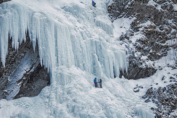 Valdez, Alaska, USA:  February 12, 2015: Two insignificant climbers stand on an ice shelf.  With gear and equipment ready they stand looking out at the ice flow.  Another climber can be seen further up the waterfall.  The ropes he used hang down in wait for the others to use on their accent up the ice.   On this cold day in February it was exciting to witness the climbers as they made their way up the frozen waterfall.