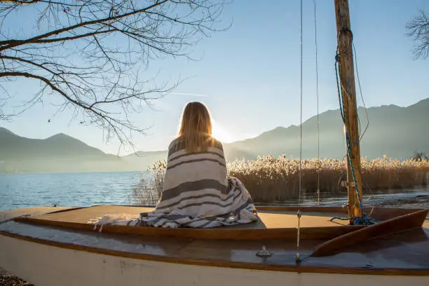 Young woman sitting on a sailboat by the lake watching sunset and relaxing.