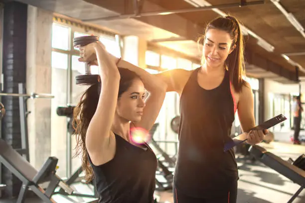 Sporty girl doing weight exercises with assistance of her personal trainer at gym.