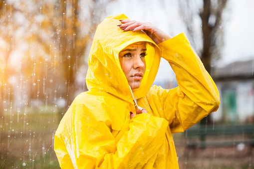 Beautiful young blonde woman standing on the rain without umbrella. She have a waterproof yellow raincoat. She looks sad and worry because it's rainy day and water is everywhere.