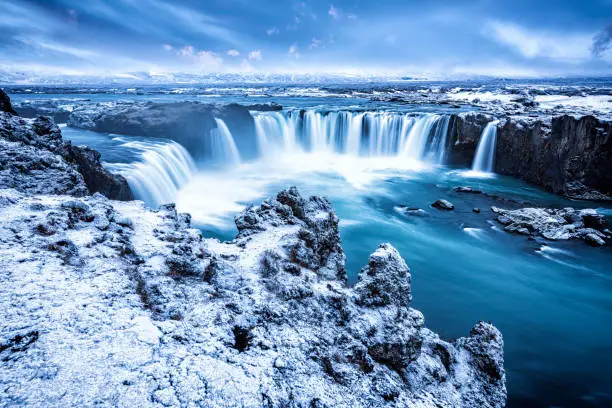 Godafoss Falls during Sunrise shows the water pouring over the edge and kicking up a misty cloud over the water with snow covering the cliffs.