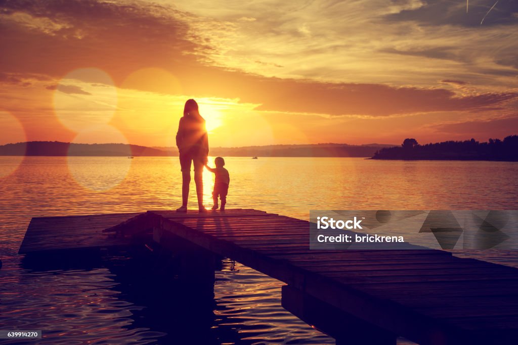 Silhouettes of Mother and Her Son by the Lake Silhouettes of Young Mother and Her Little Baby Standing on the Pier and Watching Sunset by the Lake. Happy Family Lifestyle. Motherhood and Childhood. Mom and Son. Toned Photo with Bokeh. Copy Space. Mother Stock Photo