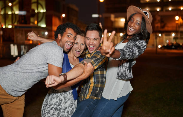 Multi-ethnic millenial group of friends taking a selfie photo Four young casual friends having fun taking pictures at an urban celebration with a cityscape view in the evening peace sign gesture photos stock pictures, royalty-free photos & images