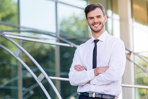Keep smiling. Confident young man keeping arms crossed and looking at camera with smile while standing outdoors with office building in the background