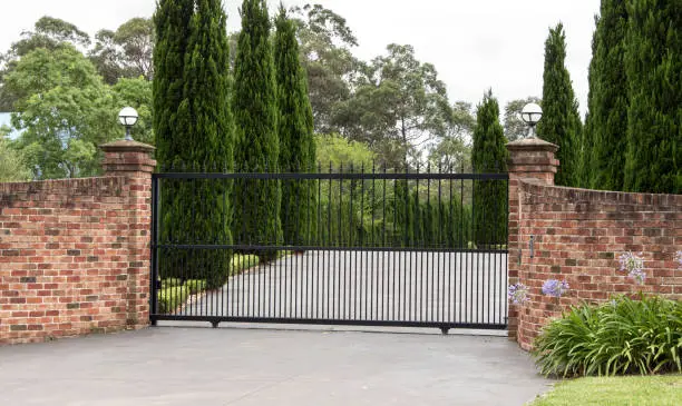 Black metal driveway entrance gates set in brick fence with pine trees in background