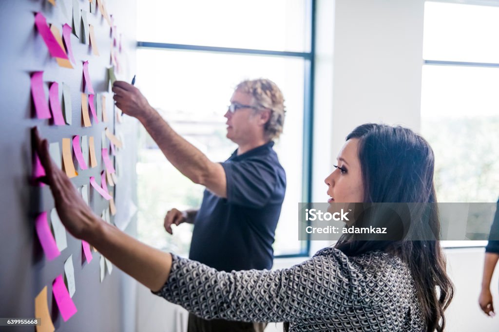 Brainstorming with Notes on the Wall An attractive young Asian businesswoman and a mature man leading a brainstorming session by putting post it notes on the wall Adhesive Note Stock Photo