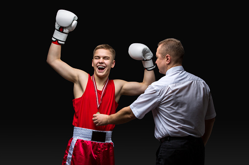 Boxing referee gives medal to young teen boxer in red form and white gloves. Winner. Studio shot on black background. Copy space.