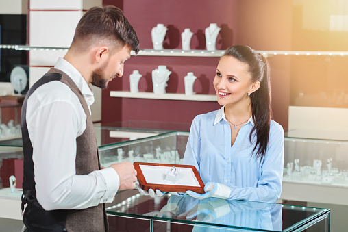 Young jeweler brunette girl holding in hands and showing for handsome man engagement ring with blue stone. Looking each other, talking and smiling. Male thinking and choosing jewelry in luxury store.