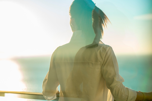 Young woman with black hair worn in a ponytaiol, wearing a white shirt and standing on a balcony enjoying a beautiful fresh morning by looking at a turquoise blue ocean with reflection of a strong morning sun. View from the back through a window glass.