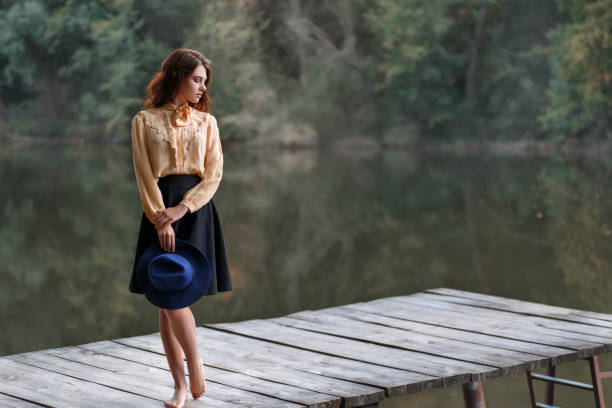 Dreaming girl on bridge with forest and river on background. Beautiful brunette woman in elegant vintage blouse with bow, skirt, floppy hat by the lake. Natural sensual caucasian woman outdoors