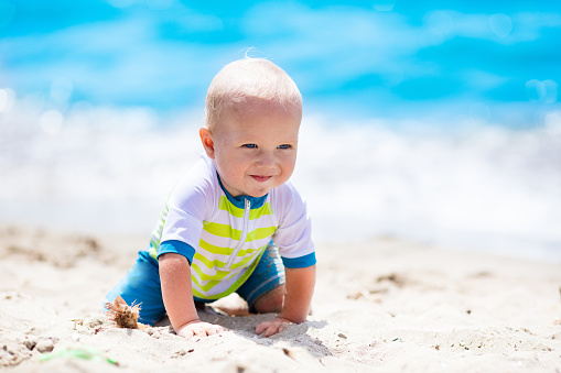 Little baby boy wearing blue rash guard suit playing on tropical ocean beach. UV and sun protection for young children. Toddler kid during family sea vacation. Summer water fun.