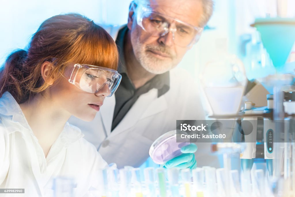 Health care professionals in lab. Attractive young female scientist and her senior male supervisor looking at the cell colony grown in the petri dish in the life science research laboratory Research Stock Photo