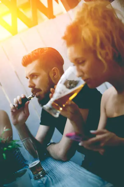 Male smoker is in focus. He is smoking electronic cigarette and looking at his friends. He has half empty glass of beer in front of him. His friend is also drinking beer while trying to see message on her phone.