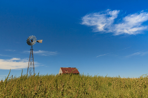 A photograph of a landscape in Kansas, USA, showing a wind turbine and small hut set on a gentle hill with a blue sky behind.