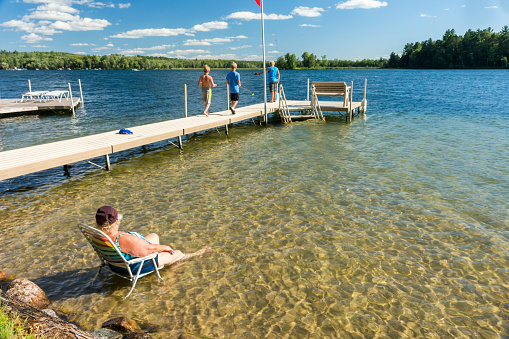 Caucasian grandmother sits in shallow water in a lounge chair watching three grandsons fishing from the dock on a summer family vacation at the lake, Hackensac, Minnesota, USA