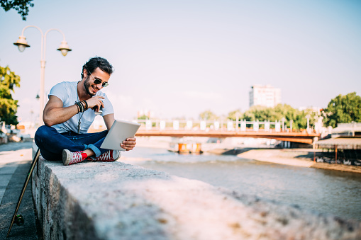 Staying ahead with new technologies.Hipster sits by the river in looking at tablet.He drinks coffee.Next to him is his longboard.He enjoys a nice summer day