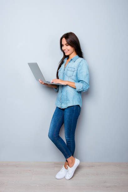 jeune femme souriante debout près du mur et tenant un ordinateur portable - human leg jeans converse shoe photos et images de collection