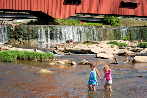 Two Caucasian girls aged 11 years wade through shallow water at the base of a covered bridge and waterfall on a hot day, Parke County, Indiana, USA