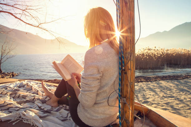 Young woman relaxing by the lake, sitting on wooden sailboat reading a book at sunset in autumn, Switzerland.