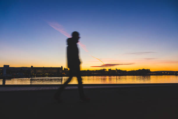 Silhouette of a man walking in Oslo at sunset. Young man walking next to the sea with some city buildings on far background. Travel and time concepts. Long exposure with blurred motion.