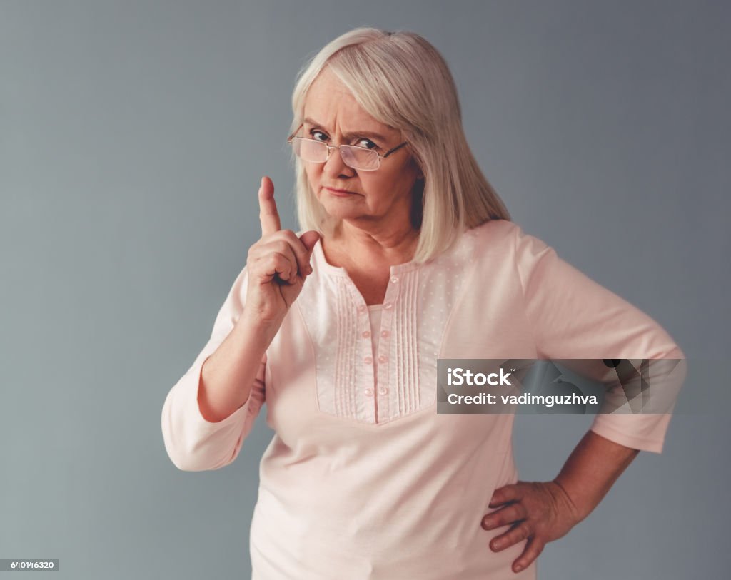 Beautiful mature woman Beautiful mature woman in eyeglasses is warning and looking at camera, on gray background Scolding Stock Photo