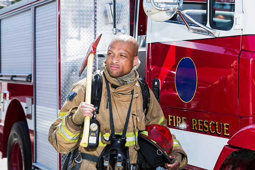 An African American fire fighter next to a fire engine, carrying an axe.  He is sweating wearing a protective suit.  He has a serious expression on his face.