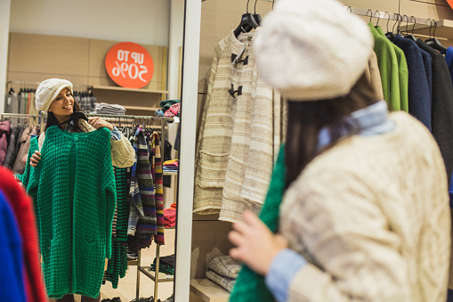 Woman holding knitted green cardigan in front of mirror in clothing store.