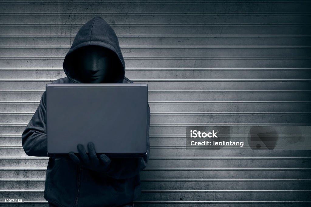 Hooded hacker with mask holding laptop while typing Hooded hacker with mask holding laptop while typing against metal roller shutter door background Hooded Shirt Stock Photo