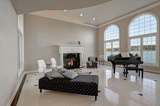 Luxury high ceiling living room features beige ivory walls framing large arched windows, traditional fireplace, black grand piano next to cozy sitting area atop glossy marble floor. Northwest, USA