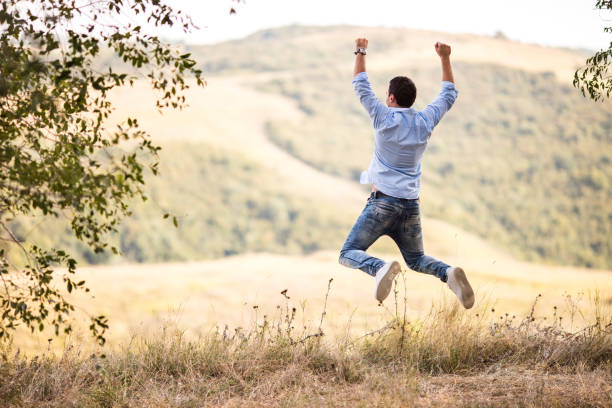 Back view of carefree man at the park jumping with arms raised.