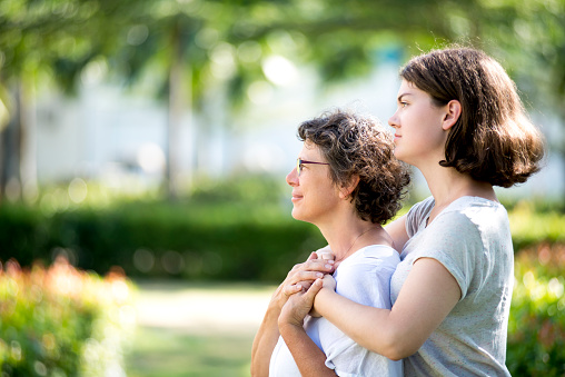 Side view of serious senior Caucasian mother wearing glasses standing embracing with her teenage daughter in park and looking away