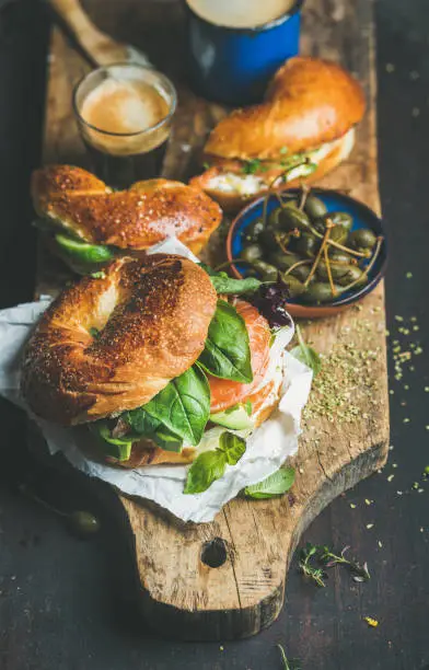 Breakfast with bagels with salmon, avocado, cream-cheese, basil, espresso coffee in glass, capers on rustic wooden board over dark scorched background, selective focus. Healthy or diet food concept