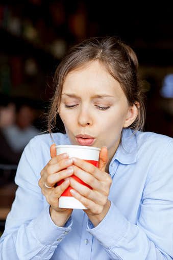 Young woman blowing on hot coffee to cool it down. Serious businesswoman drinking coffee in cafe. She holding disposable cup. Hot drink concept