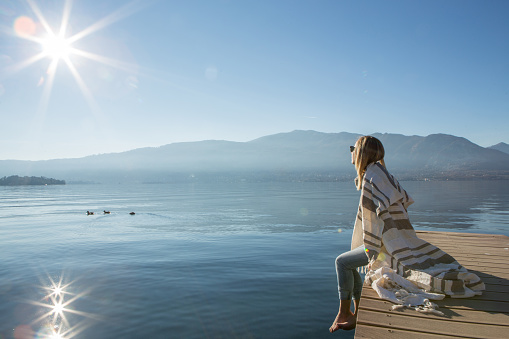 Young woman relaxes on lake pier, looks at majestic landscape. Beautiful Autumn day in Italy.