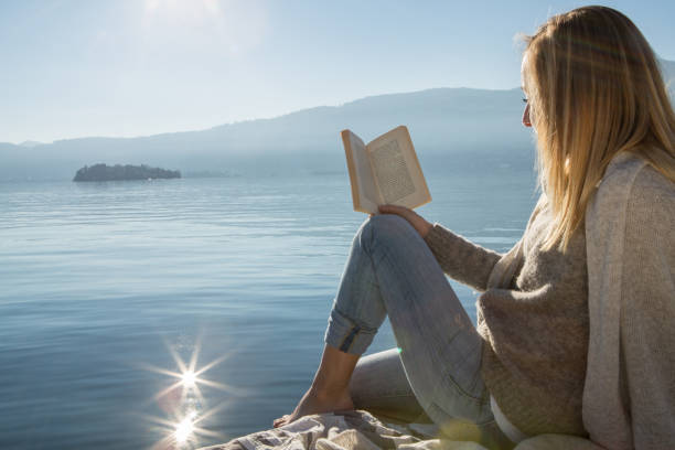 istock Young woman sits on jetty above lake, reading book 640199350