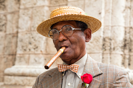 Portrait of an elegant, dark, elderly Cuban gentleman in a suit, straw boater hat, and a cigar standing in front of a wall in Havana, Cuba, 50 megapixel image.