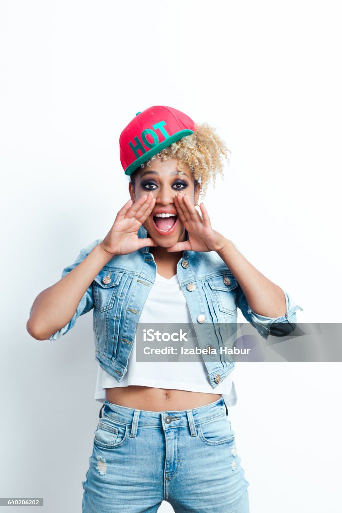 Excited afro american young woman screaming Excited afro american young woman wearing denim jacket and baseball cap, shouting at the camera. Studio shot, white background. Adult Stock Photo