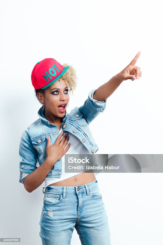 Excited afro american young woman pointing Excited afro american young woman wearing denim jacket and baseball cap, pointing with index finger at copy space. Studio shot, white background. Cool Attitude Stock Photo
